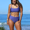 Woman wearing a purple bikini set standing on a beach with blue sky and ocean in the background.