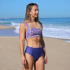 Woman in a floral bikini top and blue high-waisted bottoms standing on a beach.