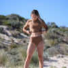 Woman in a brown bikini standing on a sandy beach with greenery in the background