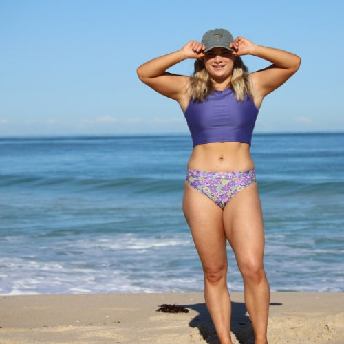 Woman in a purple bikini and cap standing on a beach with ocean in the background