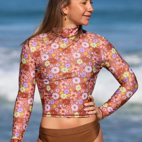 Woman wearing a floral long-sleeve top and brown shorts with a blurred ocean background