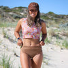 Woman in a floral crop top and beige bikini bottom standing on sandy dunes with grass and clear sky.