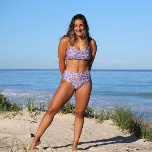 Woman in a purple floral bikini standing on a sandy beach with ocean view