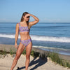 Woman in a purple floral bikini standing on a beach with ocean waves in the background