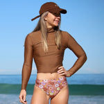 Woman in brown long-sleeve top and floral bikini bottom standing on a beach.
