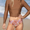 Person wearing a brown long-sleeve top and floral-patterned swimsuit on a beach.