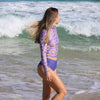 Woman in a purple floral long-sleeve rash guard and blue shorts standing on a beach with waves in the background.