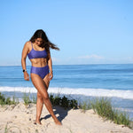 Woman in a purple bikini standing on a sandy beach with ocean in the background