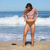 Woman in a floral long-sleeve rash guard and brown bikini bottom standing on a beach with ocean waves in the background.
