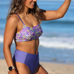 Woman wearing a purple floral bikini top and blue high-waisted bottoms on a beach.