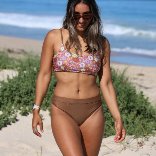 Woman in a floral bikini top and brown bottoms walking on a beach.