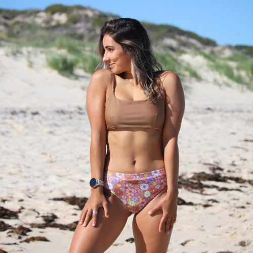 Woman in a beige bikini top and colorful floral bottom standing on a sandy beach.