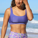 Woman wearing a purple sports bra and floral shorts on a beach.