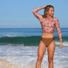 Woman in a floral long-sleeve top and brown bikini bottom standing on a beach.