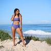 Woman in a purple bikini standing on a sandy beach with ocean waves in the background