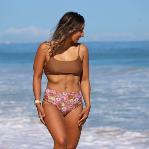 Woman in a brown bikini top and floral bottom standing on a beach with ocean in the background