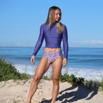 Woman in a purple swimsuit standing on a sandy beach with ocean in the background