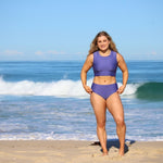Woman in a purple swimsuit standing on a beach with ocean waves in the background