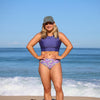 Woman in a purple bikini with floral pattern standing on a beach.