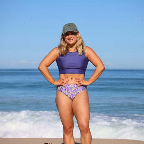 Woman in a purple bikini with floral pattern standing on a beach.